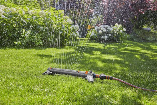 Grijze zwenksproeier spuit water op een groen gazon met bloeiende planten.