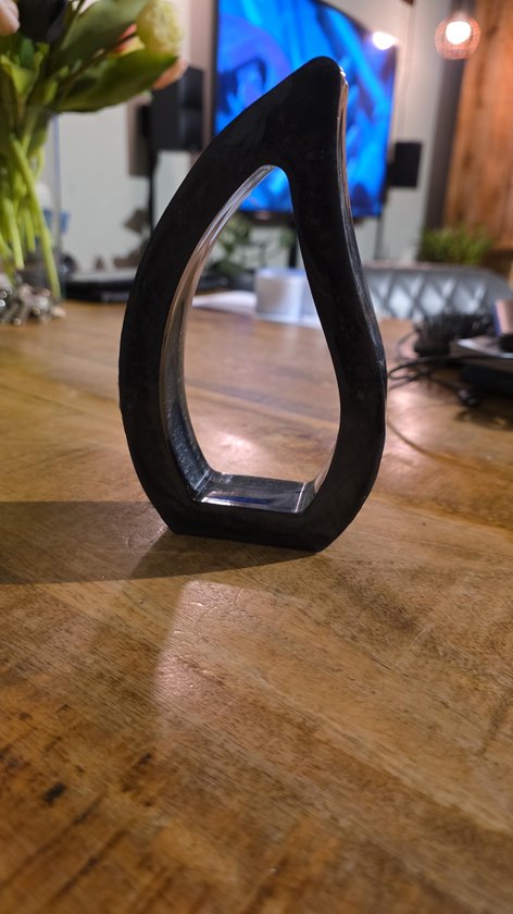 Zwarte, druppelvormige epoxy decoratie met glanzende afwerking, staand op een houten tafel met bloemen op de achtergrond.