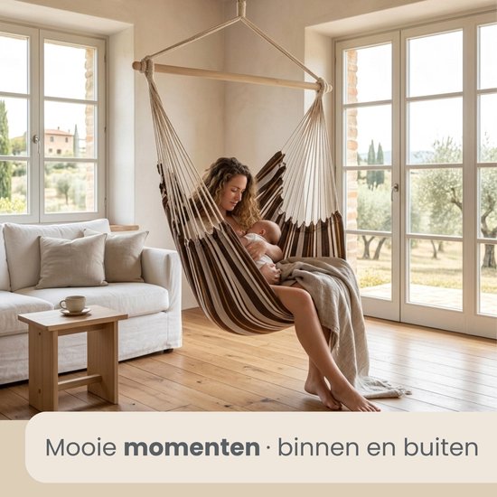 Vrouw met baby in gestreepte hangstoel, zittend in een woonkamer met uitzicht op een tuin.