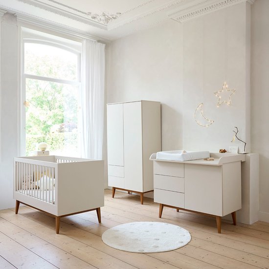 Lichtgekleurde babykamer met commode, ledikant, kledingkast en een ronde mat.