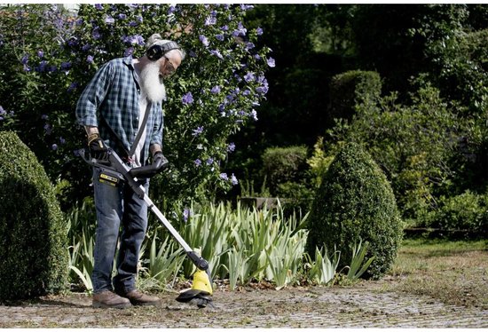 Man met baard gebruikt de gele voegenborstel in een tuin met struiken en bloemen.