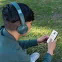 Jonge man met groene JLab GO Lux ANC koptelefoon op en telefoon in hand, zittend in gras.