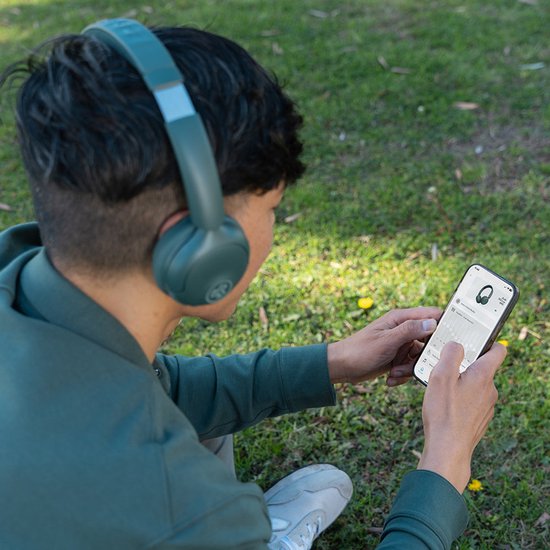 Jonge man met groene JLab GO Lux ANC koptelefoon op en telefoon in hand, zittend in gras.