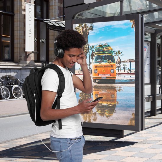 Jonge man met zwarte rugzak en koptelefoon kijkt op zijn telefoon bij een bushokje met een poster van een oranje camper.
