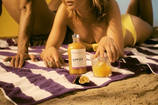 Een stel ligt op het strand met GIMBER, een glas en passievruchten.