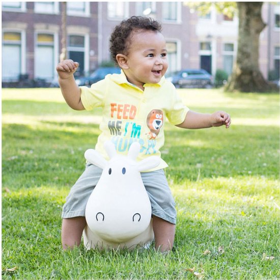 Een jong kind met krullend haar en een geel shirt zit lachend op een witte skippier koe in een grasveld.