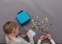 Twee kinderen bouwen met Plus-Plus speelgoedblokjes op een wit pegboard naast een turquoise koffer en verspreide blokjes op een grijze ondergrond.