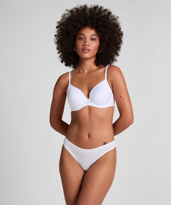 Model draagt een witte, voorgevormde beha en bijpassende slip. Ze heeft krullend haar en staat met haar handen achter haar rug.