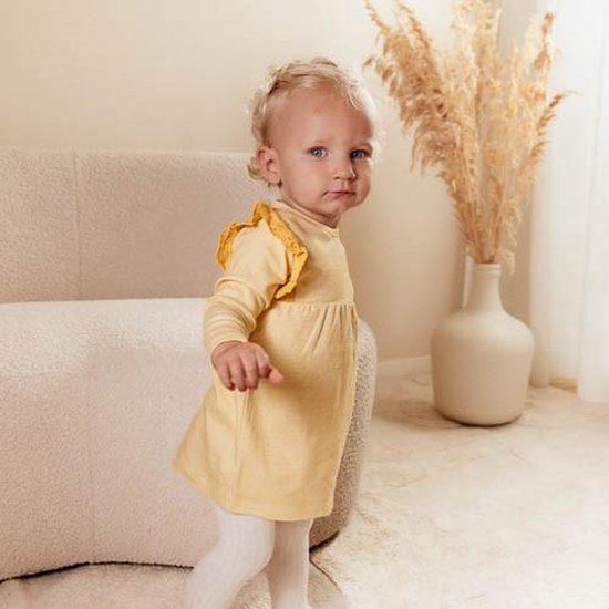 Baby met blonde krulletjes draagt een geel geruit jurkje met kanten schouderrandjes.
