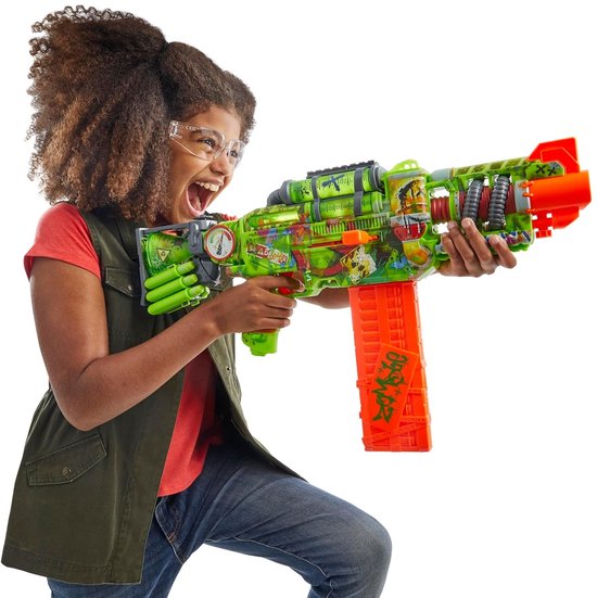 Meisje met krullend haar in groen-bruin vest, rode shirt, houdt een felgekleurde Nerf zombie-blaster vast met oranje magazijn. Ze draagt een veiligheidsbril en heeft een blije uitdrukking.