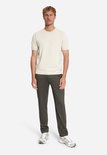 Man met een licht beige trui met korte mouwen en een donkerbruine jersey pantalon.