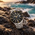Zilveren horloge met zwarte wijzerplaat en gele accenten op rotsen bij de zee.