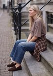 Vrouw zittend op trap met donkerblauwe wijde jeans, bruin T-shirt en geruit overhemd.