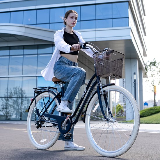 Vrouw in witte blouse en spijkerbroek op zwarte RCB-fiets met metalen mand bij een modern glazen kantoorgebouw.