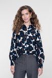 Model draagt een blouse met abstracte print in blauw, bruin en crème, gecombineerd met een grijze pantalon.