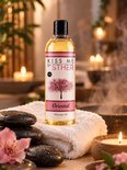 Fles 'Oriental' massageolie van Sther op een witte handdoek met zwarte stenen, bloemen en kaarsen. Met bloeiende kersenboomillustratie.