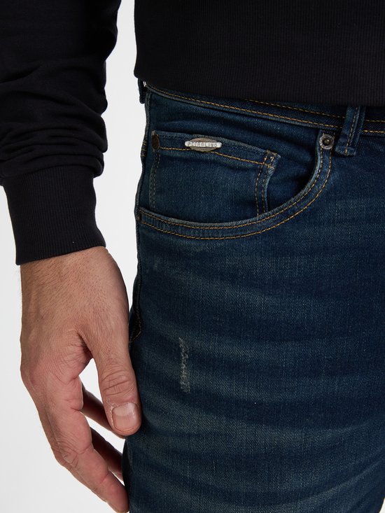 Detail van donkerblauwe spijkerbroek met stonewash en de tekst 'PETROL' op een metalen plaatje.