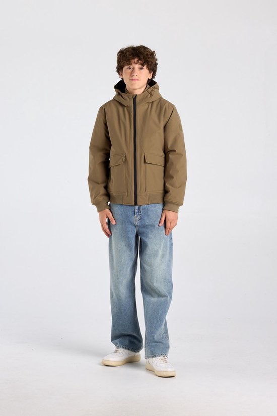 Jongen in een olijfgroene jas met capuchon, rits, zakken en lichtblauwe wijde jeans, staand op witte sneakers.