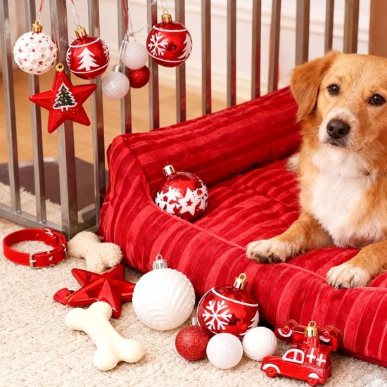 Een hond op een rood bed, omringd door kerstversieringen zoals ballen, sterren en een bot.
