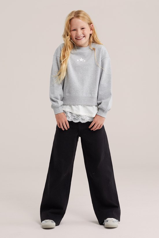 Meisje met lang blond haar draagt een grijze oversized sweater met drie witte sterren en een kanten rand, gecombineerd met een zwarte wijde broek.