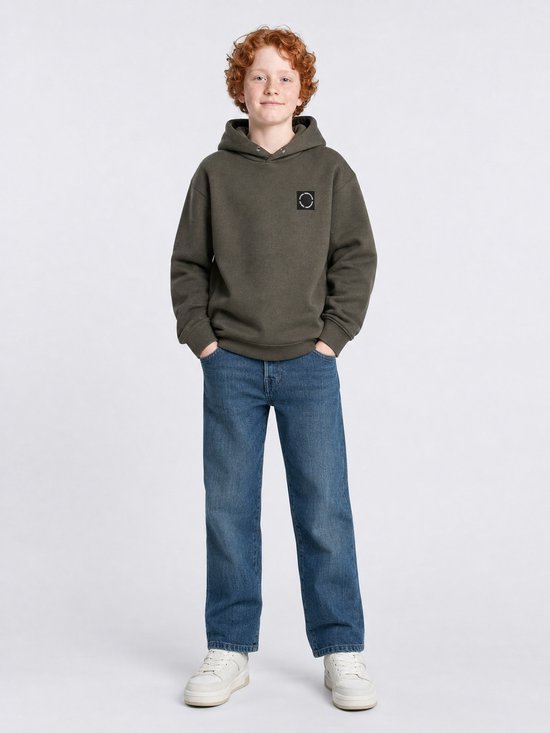Jongen met rossig haar in olijfgroene hoodie met capuchon, blauwe jeans en witte sneakers.