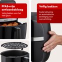Zwarte airfryer, met de mand eruit gehaald om gefrituurde snacks te tonen. Een hand raakt de zijkant aan. Tekst: "PFAS-vrije antiaanbaklaag Veilig bakken voor het hele gezin." "Veilig bakken Koel huis en automatische oververhittingsbeveiliging."