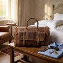 Luxe bruine leren weekendtas op een houten bankje, naast een overhemd, paspoort en zonnebril.