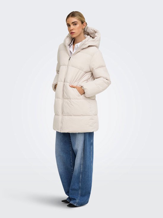 Model in een beige pufferjas met capuchon en blauwe wijde jeans, staand in profiel.