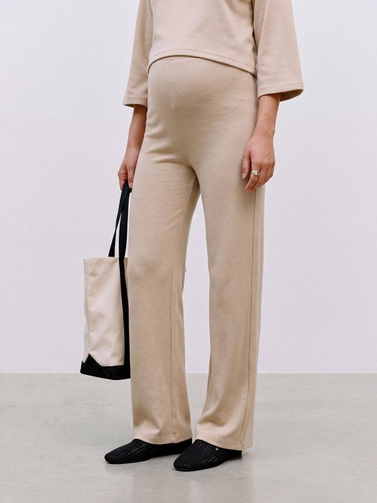 Een vrouw met zwangere buik, van de zijkant gezien, draagt een beige broek met wijde pijpen en een bijpassende top. Ze draagt een tas en zwarte schoenen.