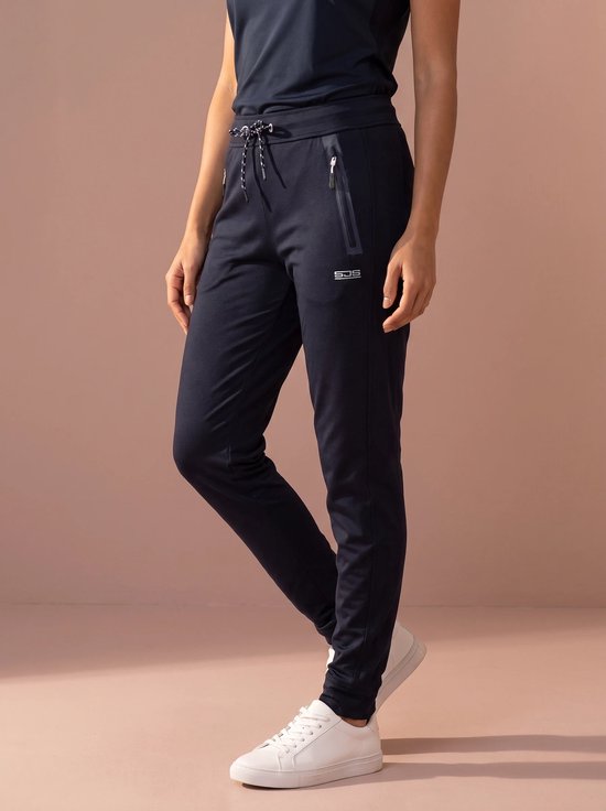 Model draagt een marineblauwe sportbroek met ritszakken en SJS-logo, gecombineerd met witte sneakers.