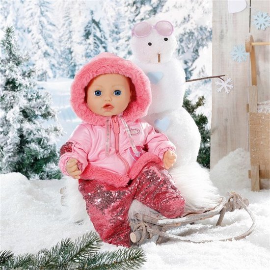 Babypop met blauwe ogen, zittend in een slee bij een sneeuwpop. Draagt een roze winterjas met bontkraag en glitterbroek.