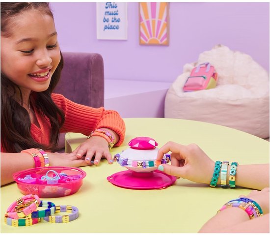 Meisje met oranje trui maakt armbandjes met de roze Pop Style armbandjesmaker, met accessoires op tafel.