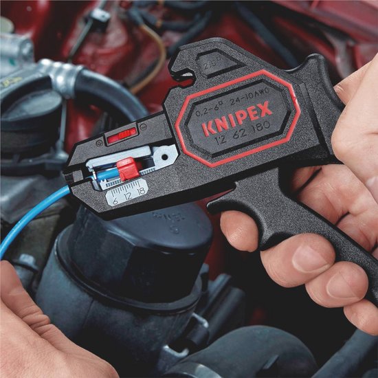 Een hand houdt een zwarte Knipex kabelstripper met rode accenten vast, die een blauwe kabel strippen in een motorruimte.