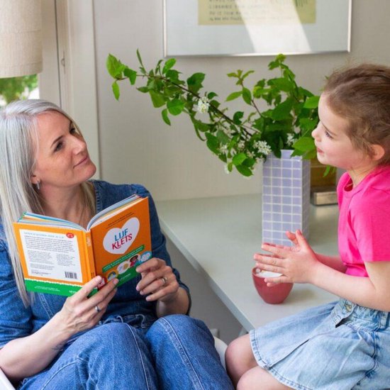 Een vrouw met grijs haar in spijkerbroek leest een boek 'Lijfklets' aan een jong meisje in een roze shirt en spijkerrok. Ze zitten aan een tafel met een vaas met bloemen.