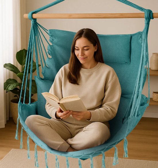 Vrouw in beige kleding leest een boek in een turquoise hangstoel met franjes, kussens en houten spreidstok.