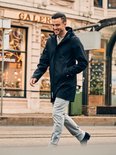 Man loopt buiten in een zwarte jas, grijze jeans, lachend.