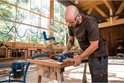 Man met veiligheidsbril en gehoorbescherming gebruikt een Bosch multitool aan een houten tafel, met een stofafzuigsysteem aangesloten.