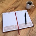 Een open rood dagboek met een pen op de rechterpagina en een mok koffie op een houten tafel.