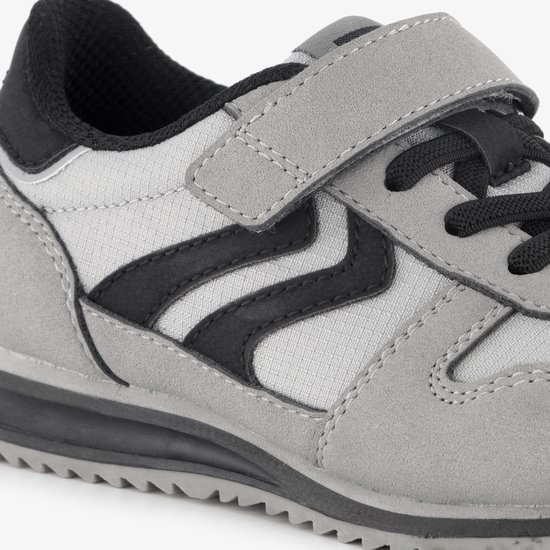 Detail van een grijze sneaker met klittenbandsluiting, zwarte zigzagstrepen en gestikte details.