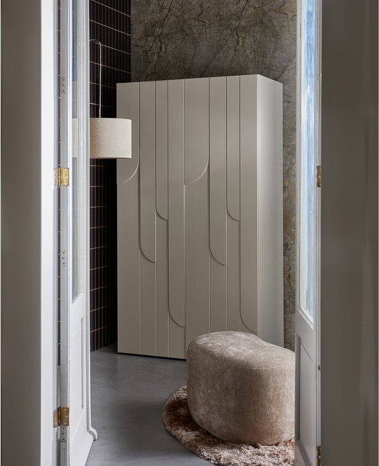 Een naturel fluwelen poef staat op een rond vloerkleed voor een moderne kledingkast.