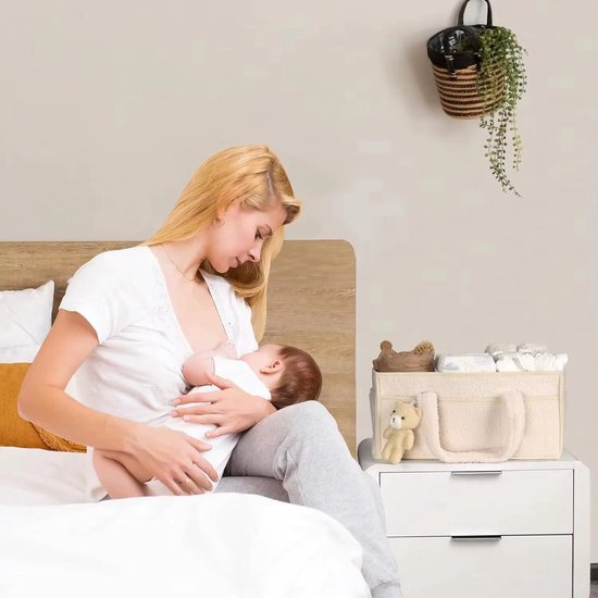 Vrouw voedt baby op bed, met een beige commandomand naast zich, gevuld met babyartikelen.