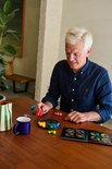Een oudere man met een blauw shirt speelt het IQ Circle spel aan een houten tafel. Hij houdt een rood stuk vast en kijkt naar het boek.
