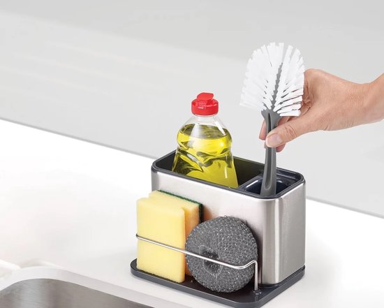 Une main place une brosse verte dans un organisateur d'évier en acier inoxydable avec éponge et savon.