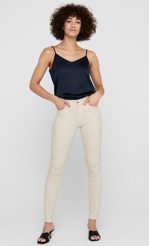 Model in crème skinny jeans met zwarte top met spaghettibandjes en sandalen.