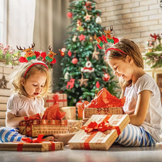 Twee kinderen met kerstdiademen met rendieren, zittend bij een kerstboom en cadeaus.