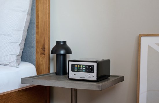 Zwarte Sonoro radio met zilverkleurig display en bedieningspaneel, staand op een betonnen nachtkastje naast een bed.