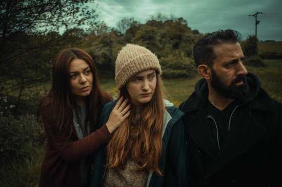 Drie personen staan buiten, twee vrouwen en een man. Een vrouw legt een hand op de schouder van een jonger meisje.