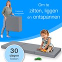 Vrouw houdt opgevouwen grijze matras met handgrepen vast. Kind speelt op matras.