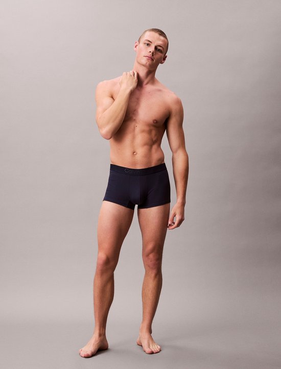 Man met gespierd bovenlichaam en armen gekruist, staand in donkerblauwe Calvin Klein boxershort.