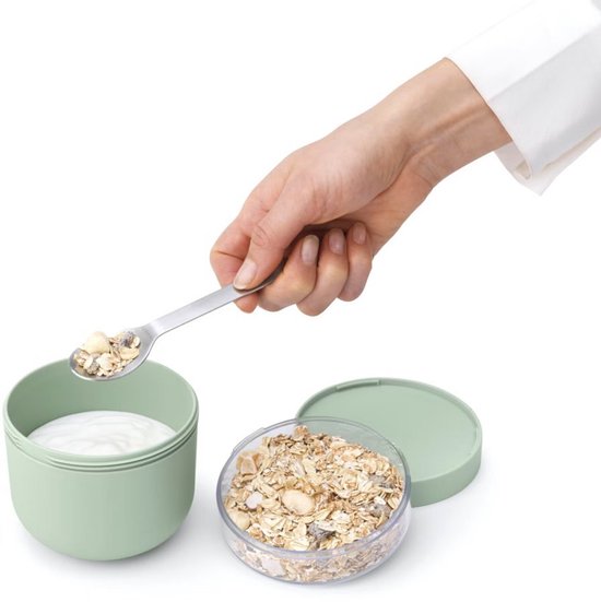 Hand vult een lepel met muesli boven een lichtgroene beker met yoghurt.